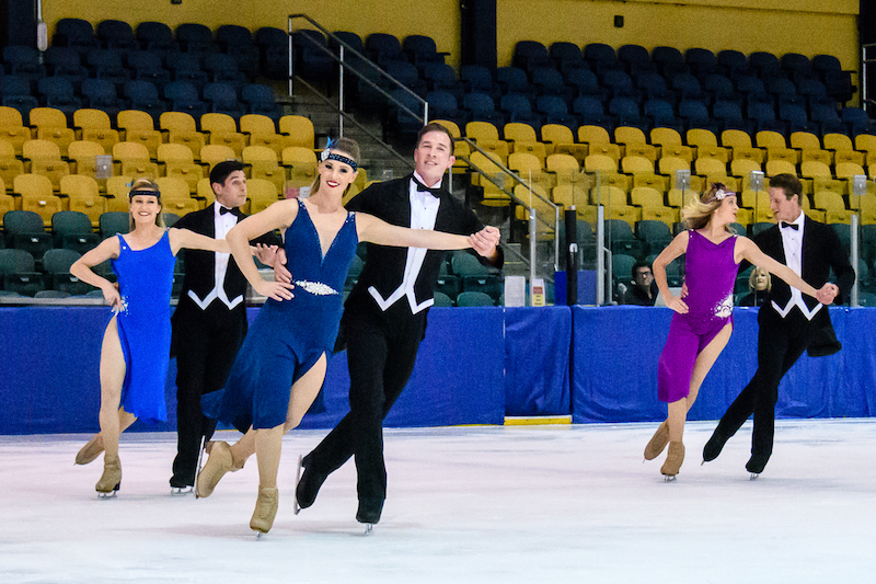 Three couples, the women in flapper-inspired blue dresses and the men in tuxes, skate hand in hand with their partners